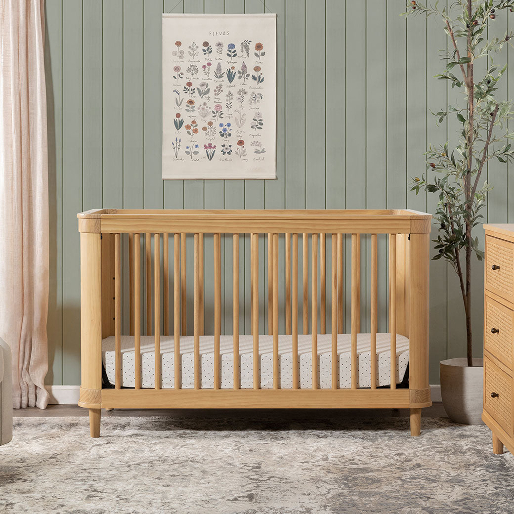 Front view of The Namesake Marin 3-in-1 Convertible Crib next to a plant and dresser in -- Color_Honey/Honey Cane