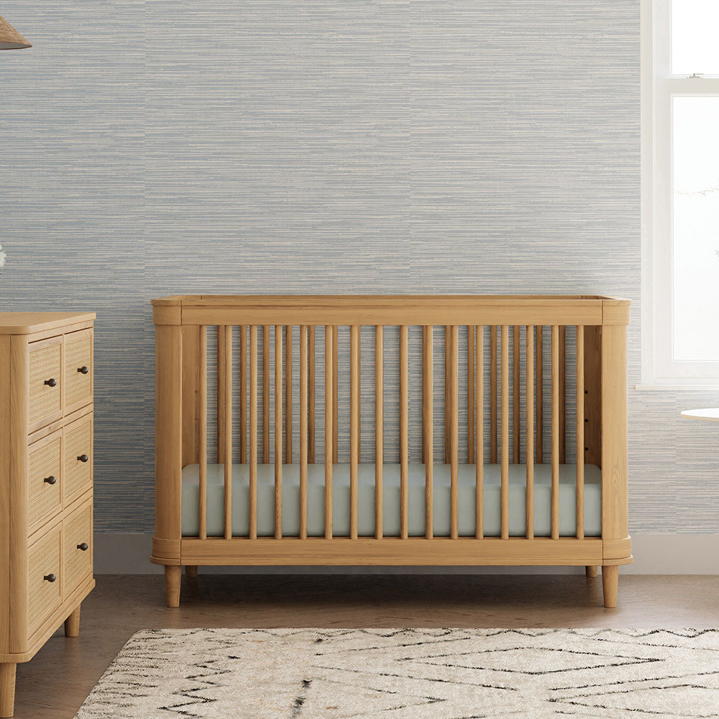 The Namesake Marin 3-in-1 Convertible Crib next to a dresser in -- Color_Honey/Honey Cane