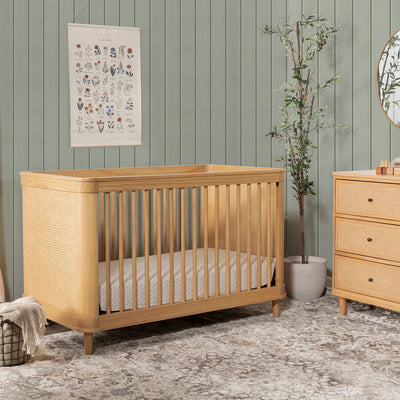 The Namesake Marin 3-in-1 Convertible Crib in a green room next to a plant and dresser  in -- Color_Honey/Honey Cane