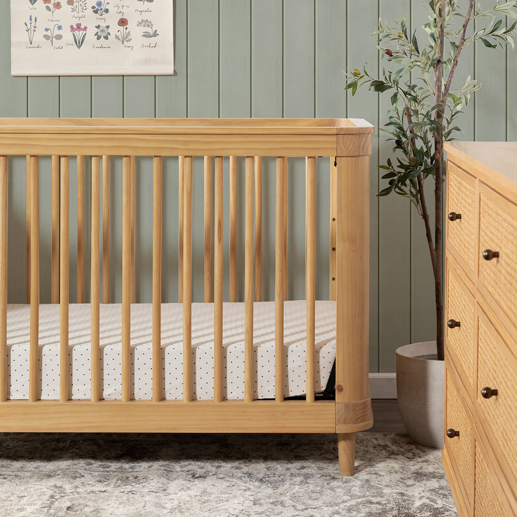 Closeup of The Namesake Marin 3-in-1 Convertible Crib next to a plant and dresser in -- Color_Honey/Honey Cane