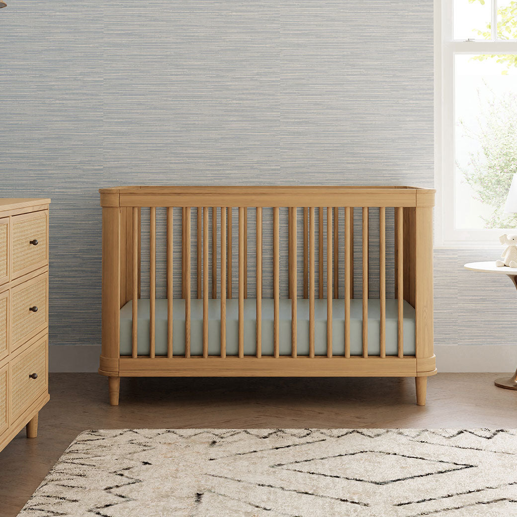 Front view of The Namesake Marin 3-in-1 Convertible Crib next to a window and dresser in -- Color_Honey/Honey Cane