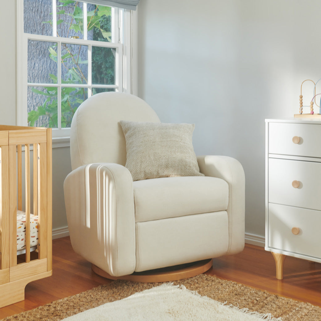 The Babyletto Nami Glider Recliner with a pillow on it next to a crib and dresser  in -- Color_Performance Cream Eco-Weave With Light Base
