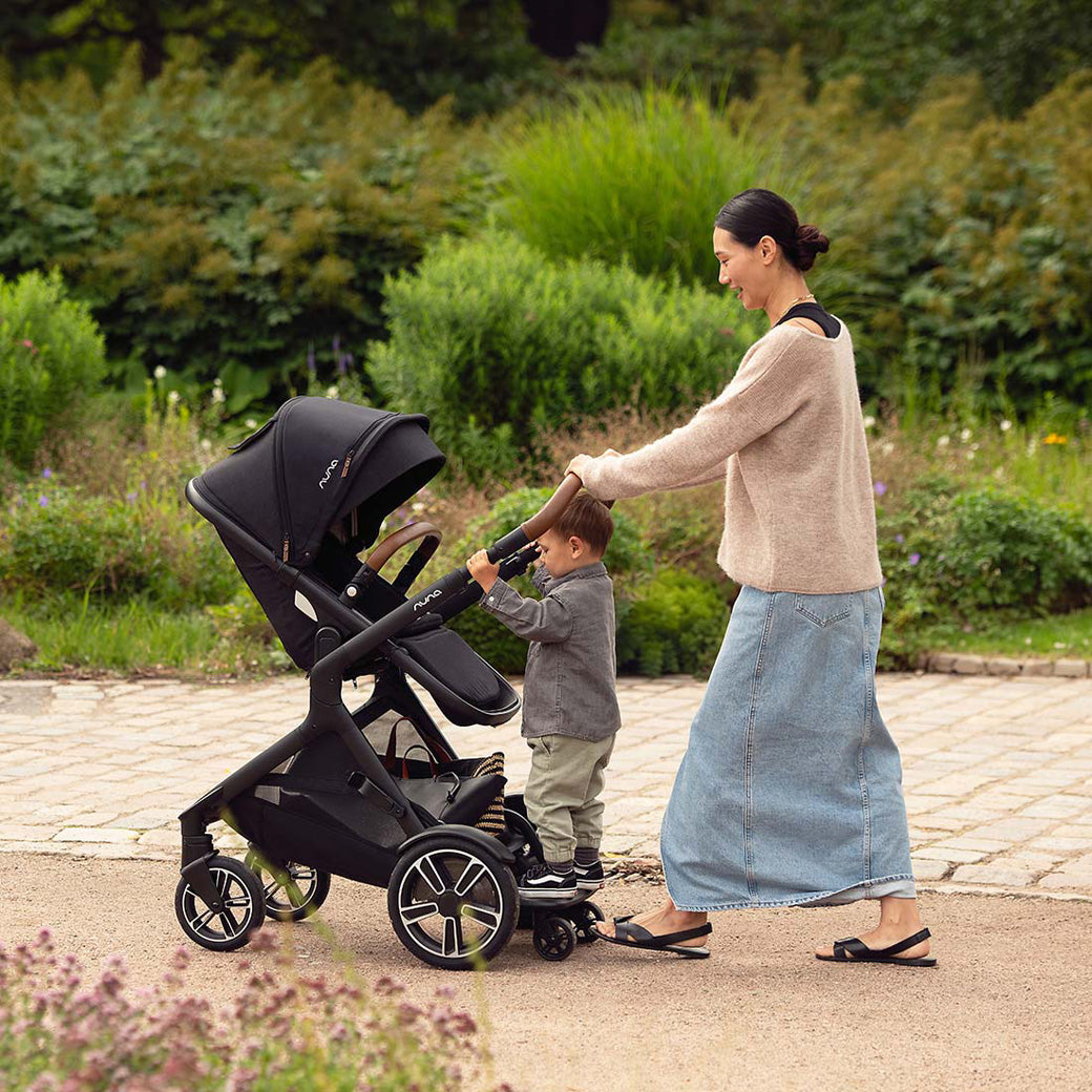 Mom pushing a child that is standing on the ride-along board of the Nuna DEMI Next Stroller in -- Color_Caviar