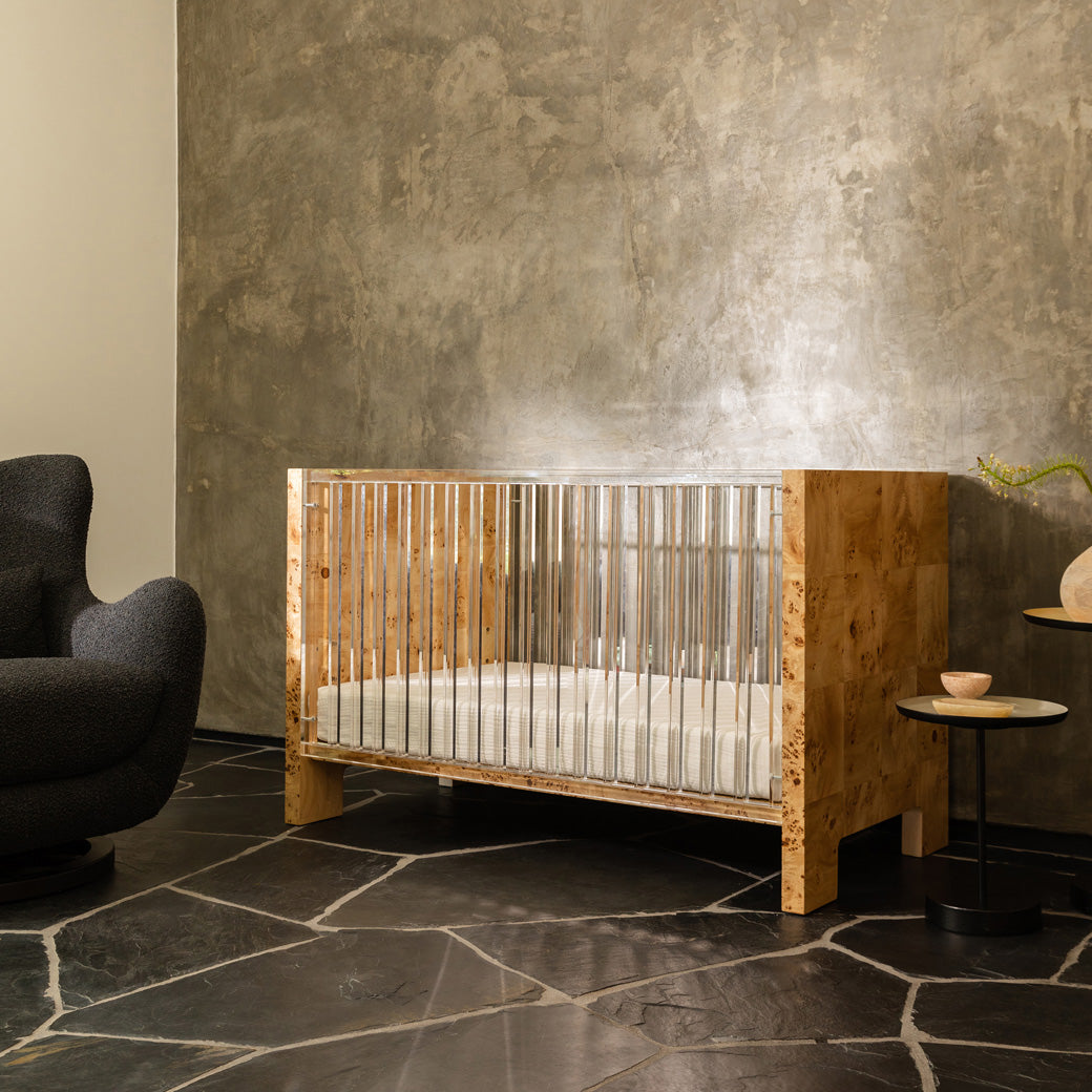 Nursery Works Altair Crib next to a recliner and coffee table in -- Color_Clear Acrylic with Burl
