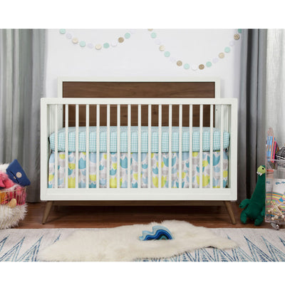 Front view of  Babyletto's Palma 4-in-1 Convertible Crib next to a basket and toys  in -- Color_Warm White with Natural Walnut