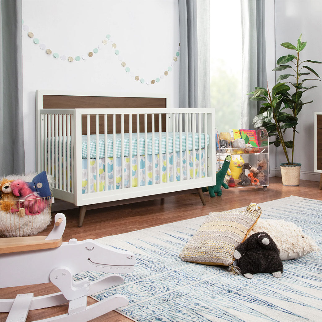  Babyletto's Palma 4-in-1 Convertible Crib  next to a basket and toy shelf in -- Color_Warm White with Natural Walnut
