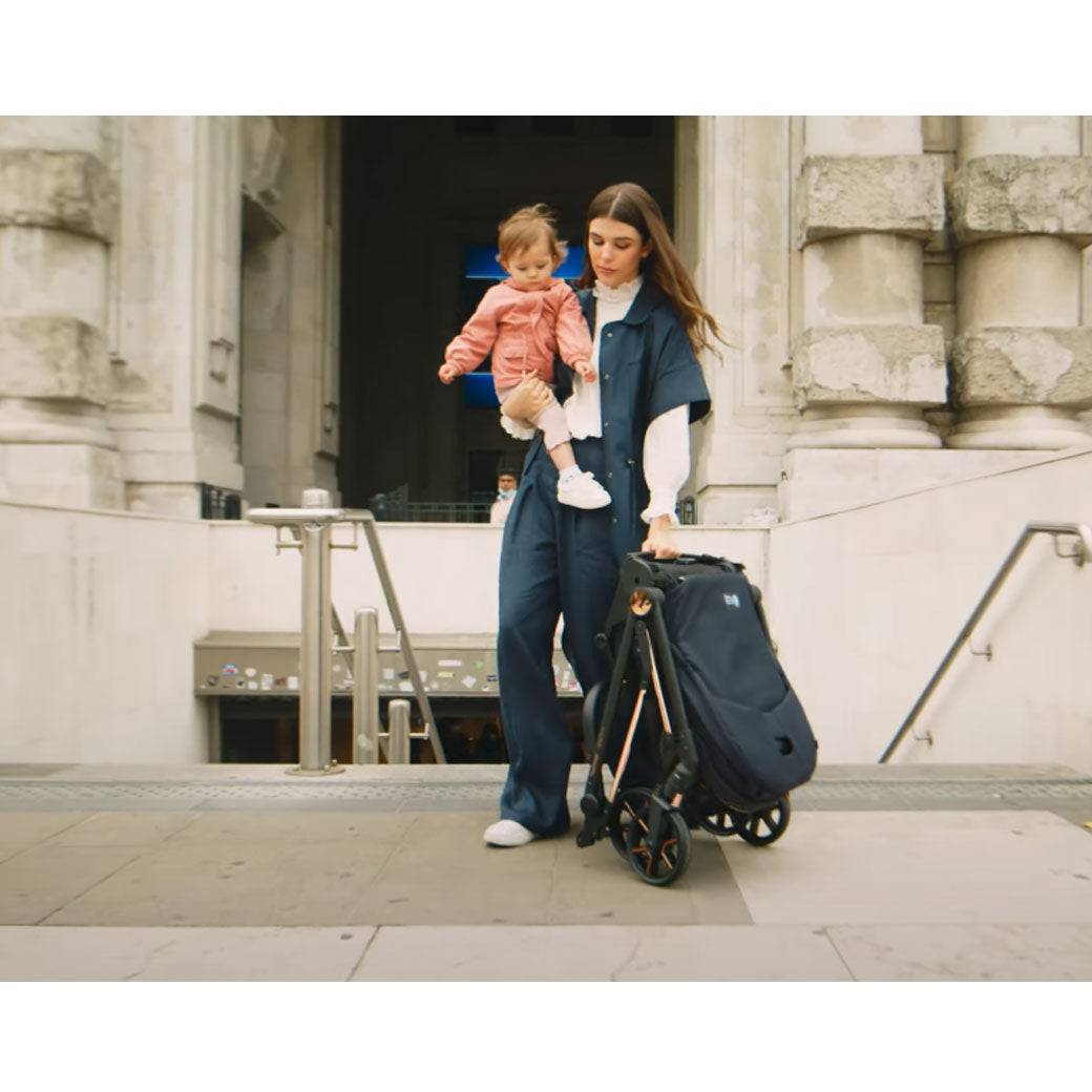 Woman holding a baby and carrying the folded Peg Perego Vivace Stroller in -- Lifestyle