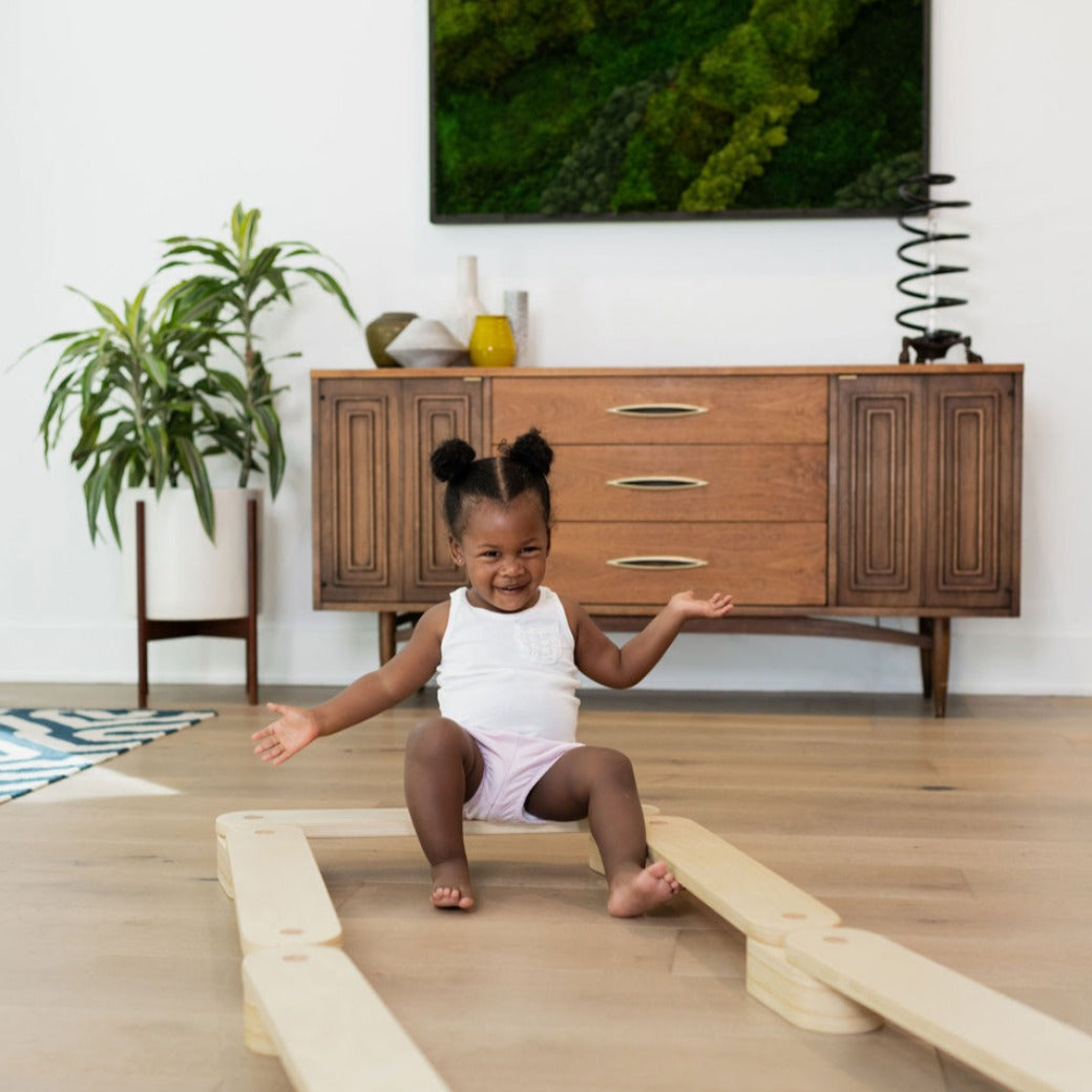 A child sitting on the Poppyseed Play Balance Beam in front of a dresser 