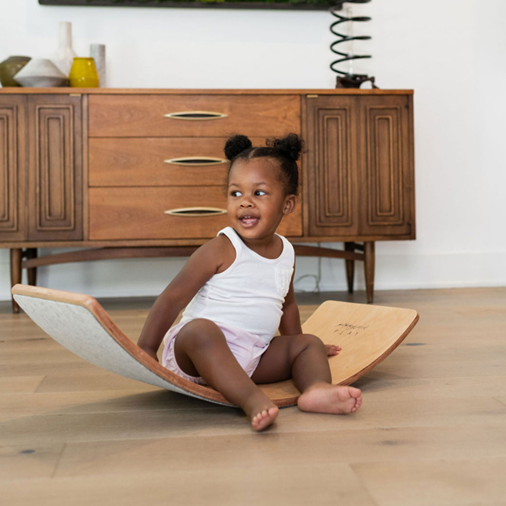 Child sitting and laughing on the Poppyseed Play Balance Board in -- Color_Gray Felt Bottom