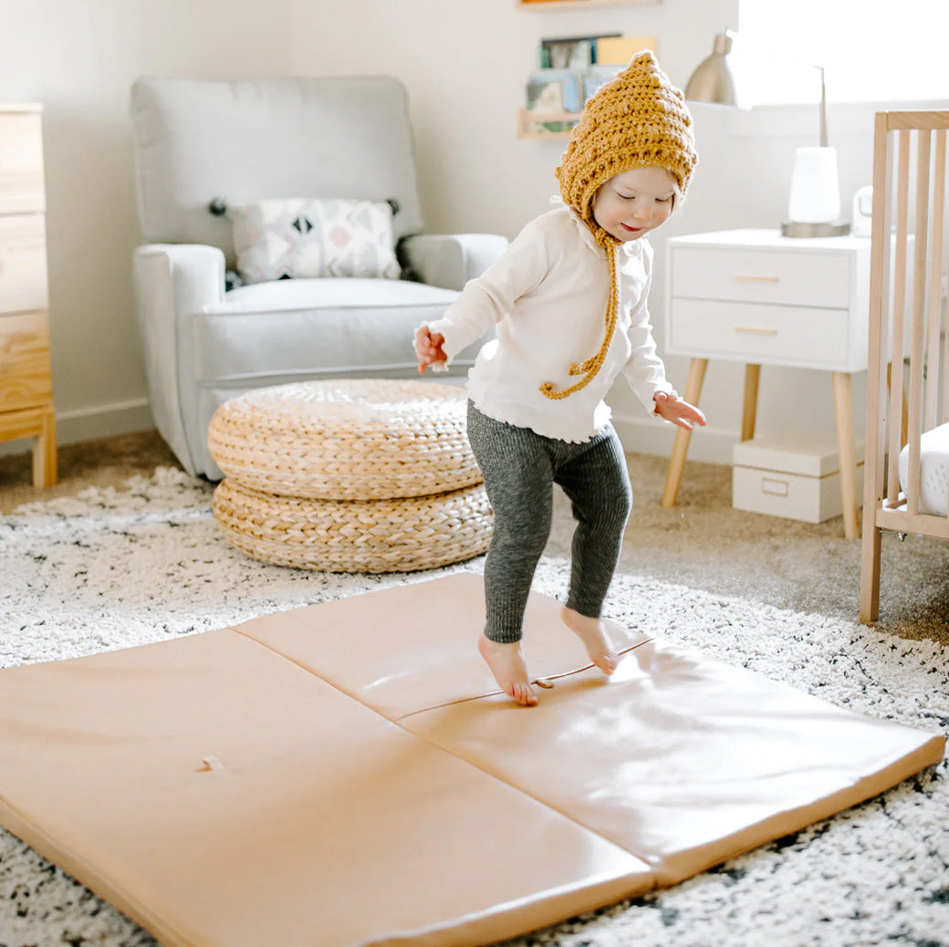 A child with a hat jumping on the Toki Mats Vegan Leather Play Mat in -- Color_Sand