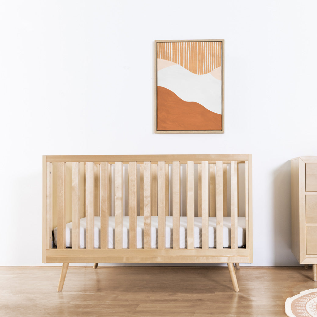 Front view of Ubabub Nifty Timber 3-In-1 Crib under a picture and next to a dresser  in -- Color_Natural Birch