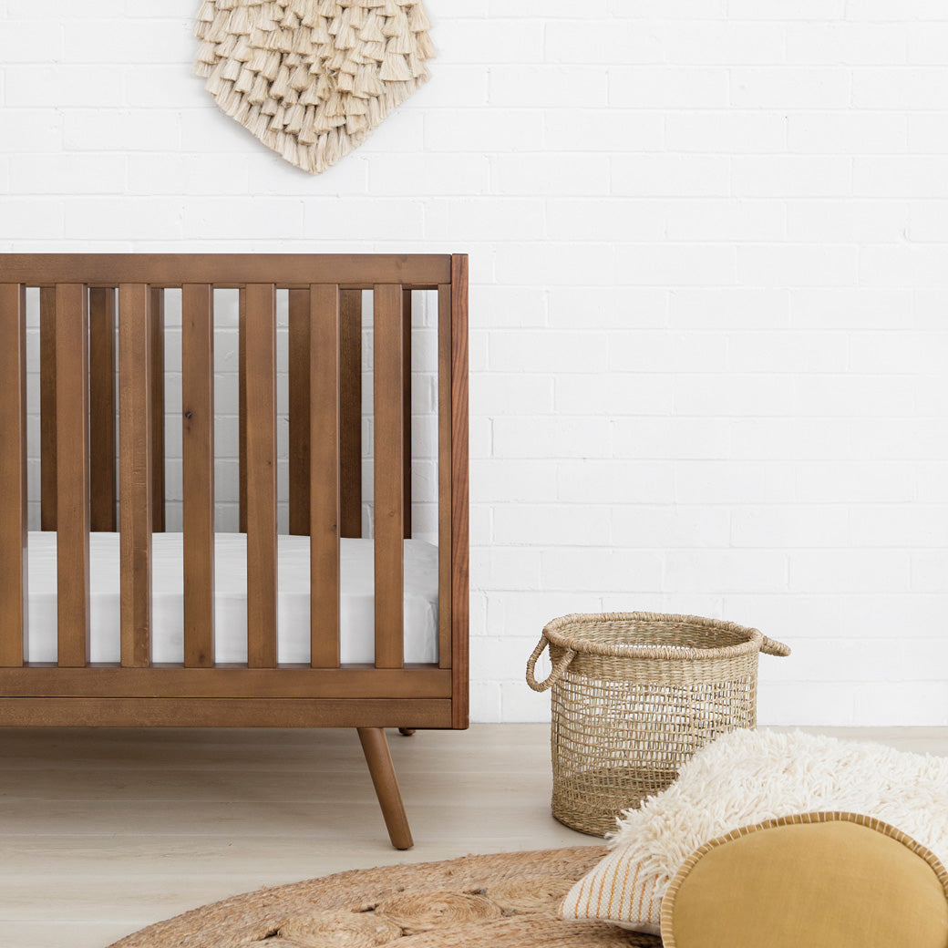 Closeup view of Ubabub Nifty Timber 3-In-1 Crib next to a basket in -- Color_Walnut