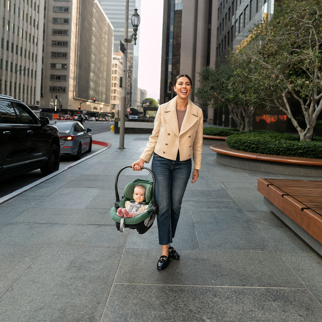 Mom walking down the street carrying baby in the Uppababy Aria Infant Car Seat in -- Color_Gwen