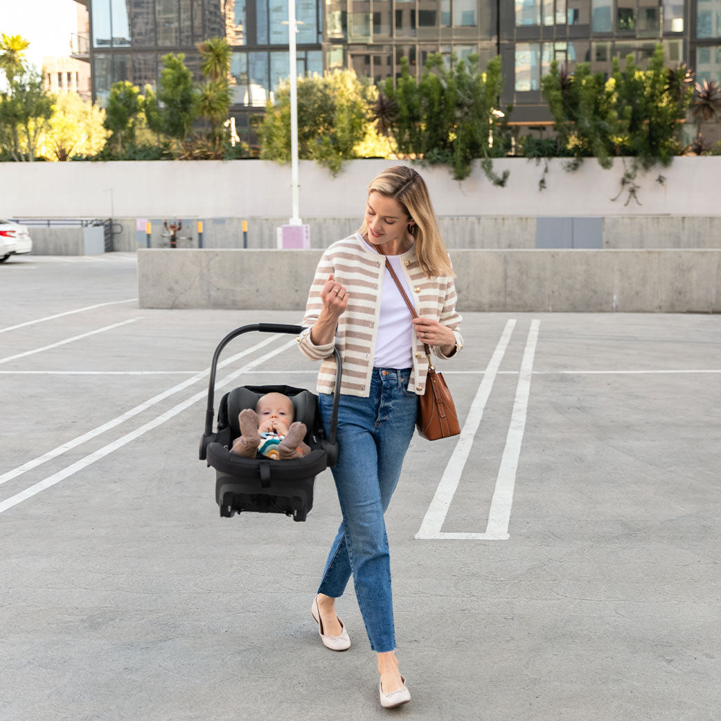Mom walking and carrying a baby in the Uppababy Aria Infant Car Seat in -- Color_Jake