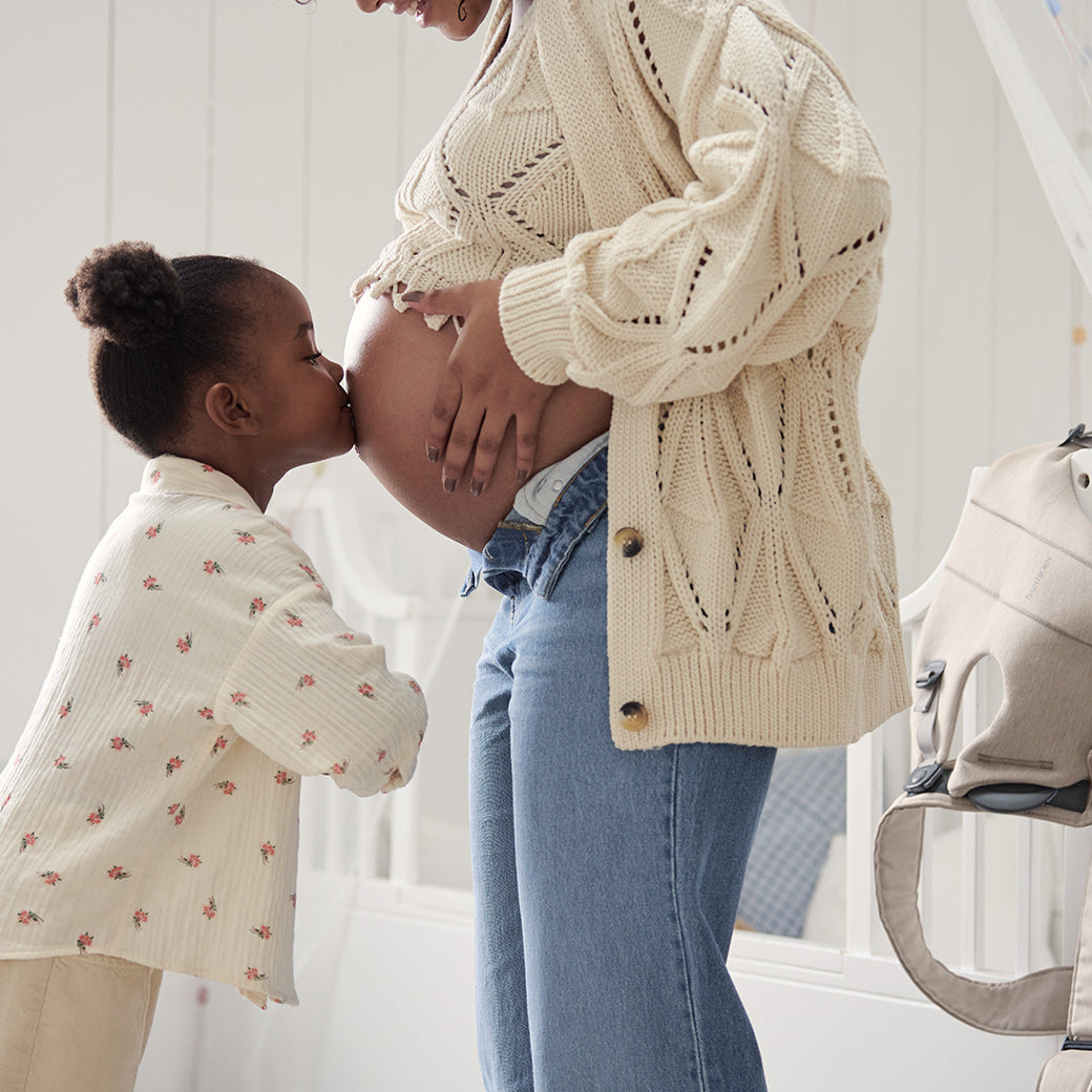 Toddler kissing mom's pregnant belly with BABYBJÖRN Baby Carrier Mini in the background in -- Color_Light Beige 3D Jersey
