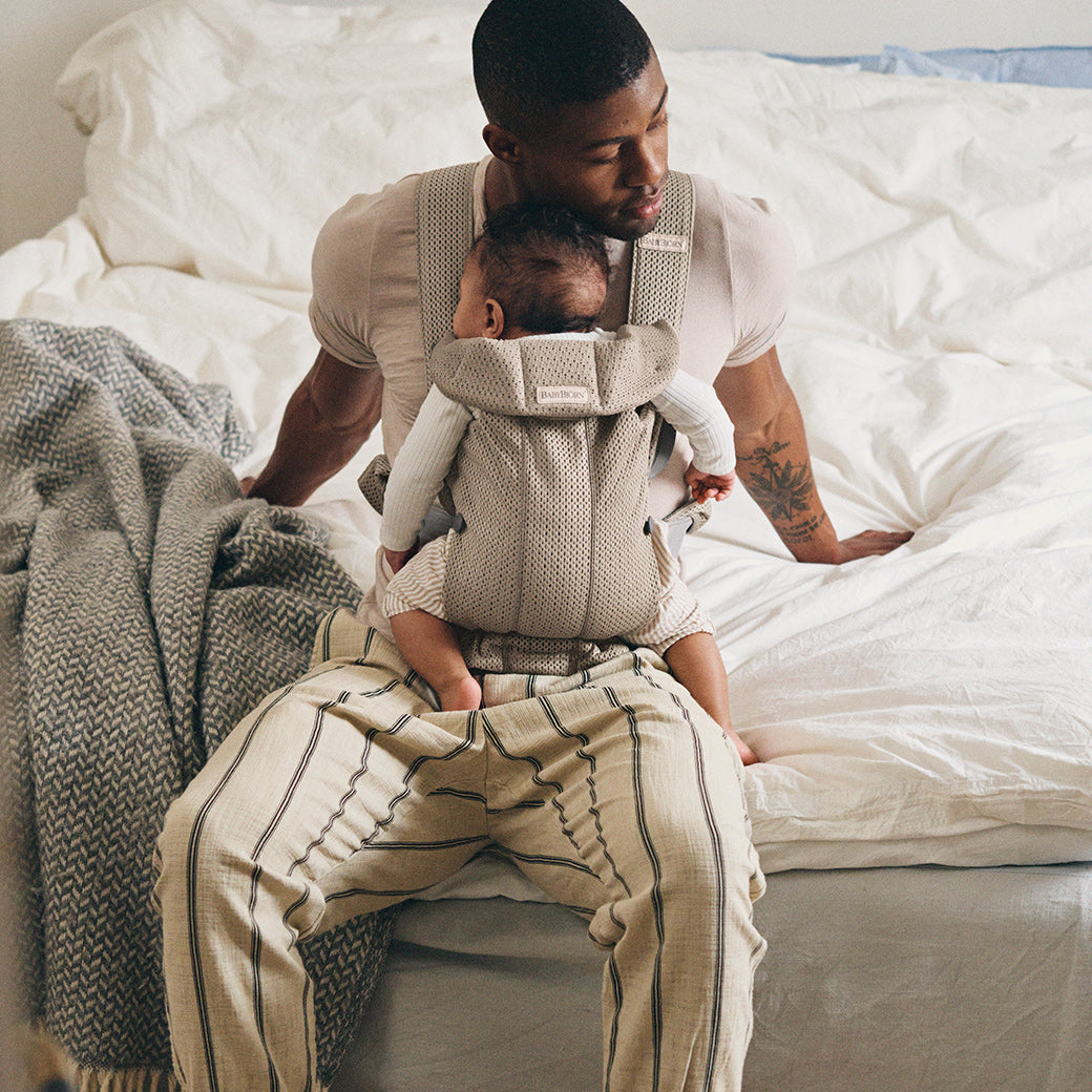 Dad sitting on bed holding baby in BABYBJÖRN Baby Carrier Mini in -- Color_Beige Grey 3D Mesh