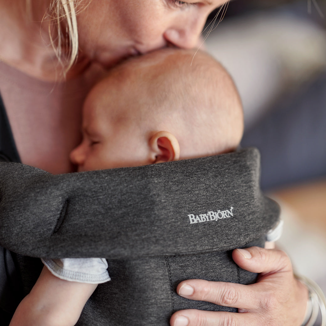 Closeup of mom kissing baby's forehead and carrying baby in the BABYBJÖRN Baby Carrier Mini in -- Color_Charcoal 3D Jersey