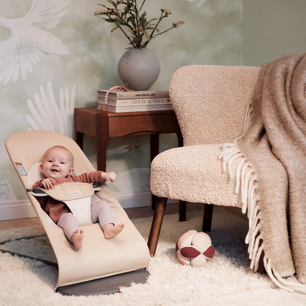Baby in BABYBJÖRN Bouncer Balance Soft next to chair and coffee table  in -- Color_Khaki/Beige Cotton With Light Grey Frame