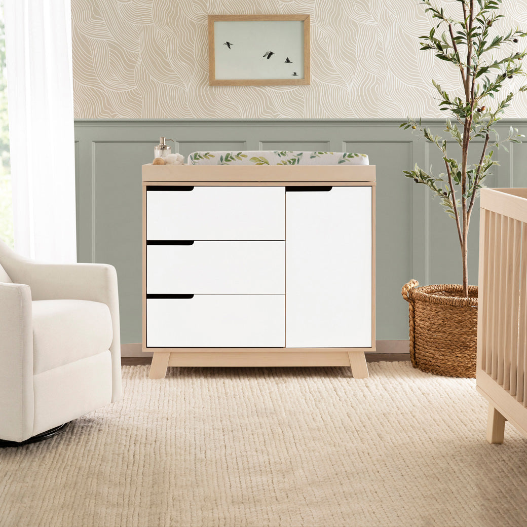 Lifestyle front view of The Babyletto Hudson Changer Dresser next to a chair in -- Color_Washed Natural/White