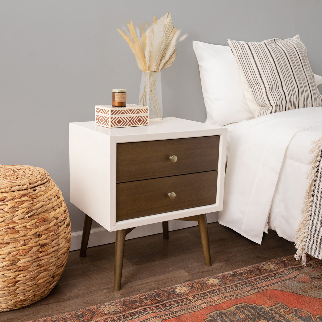 Babyletto's Palma Nightstand With USB Port next to a bed and basket in -- Color_Warm White with Natural Walnut