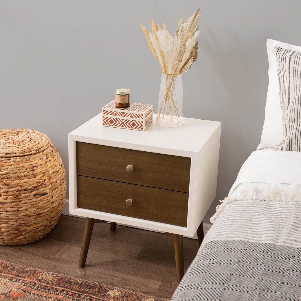 Babyletto's Palma Nightstand With USB Port next to a bed and basket  in -- Color_Warm White with Natural Walnut