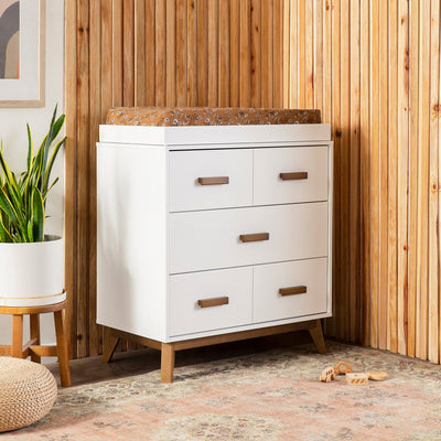 Babyletto's Scoot 3-Drawer Changer Dresser in a room next to a plant  in -- Color_White/Natural Walnut