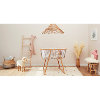 Distant profile view of Charlie Crane KUMI Cradle in Hazelnut in a baby room filled with toys -- Color_Hazelnut