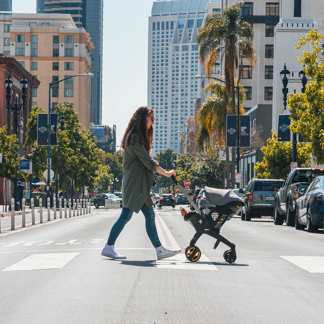 Woman walking on a crosswalk with her baby in the Doona Infant Car Seat and Stroller in -- Color_Greyhound