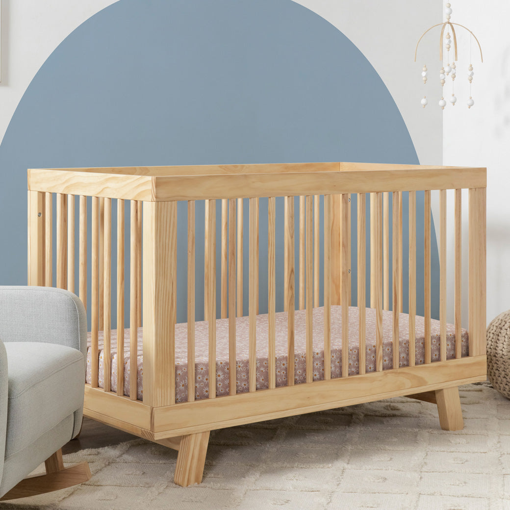 Angled view of Babyletto Hudson 3-in-1 Convertible Crib And Toddler Rail  in nursery setting with daisy sheets in -- Color_Natural