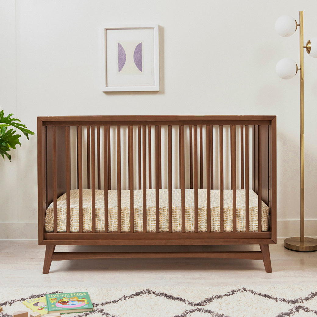 Front view of Babyletto's Peggy 3-in-1 Convertible Crib next a plant and lamp in -- Color_Natural Walnut