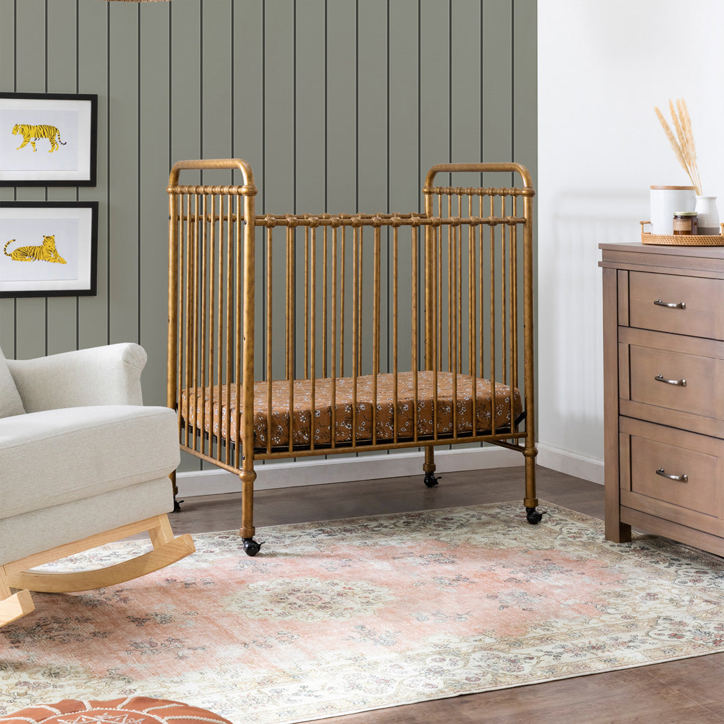 Namesake`s Abigail 3-in-1 Convertible Mini Crib next to a rocking chair in -- Color_Vintage Gold