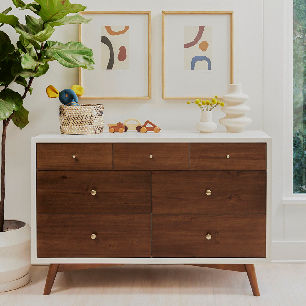 Babyletto's Palma 7-Drawer Assembled Double Dresser next to a plant in -- Color_Warm White with Natural Walnut