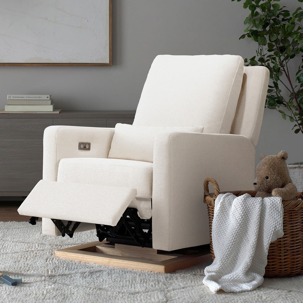 Babyletto Sigi Electronic Glider Recliner next to a basket with a blanket and a teddy bear with footrest up  in -- Color_Performance Cream Eco-Weave