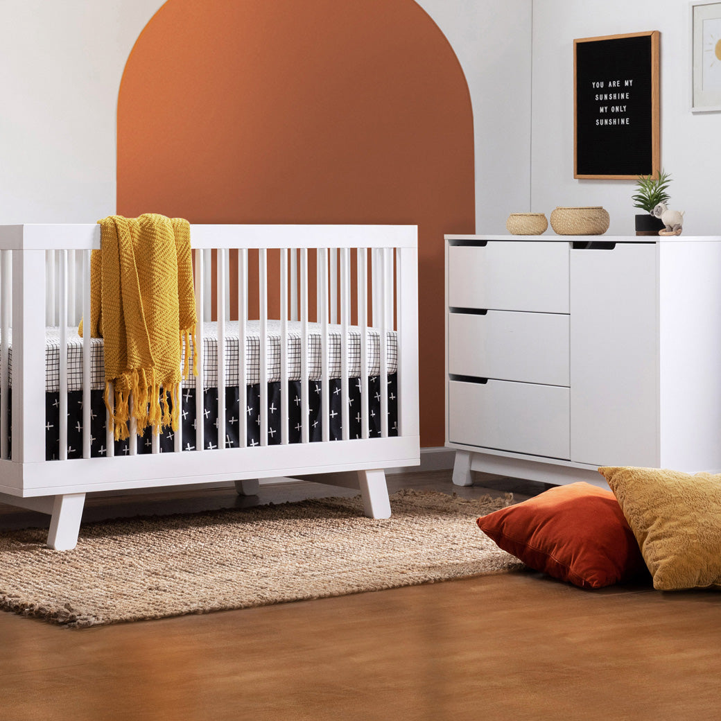 Lifestyle side view of The Babyletto Hudson Changer Dresser next to a crib in -- Color_White/Grey