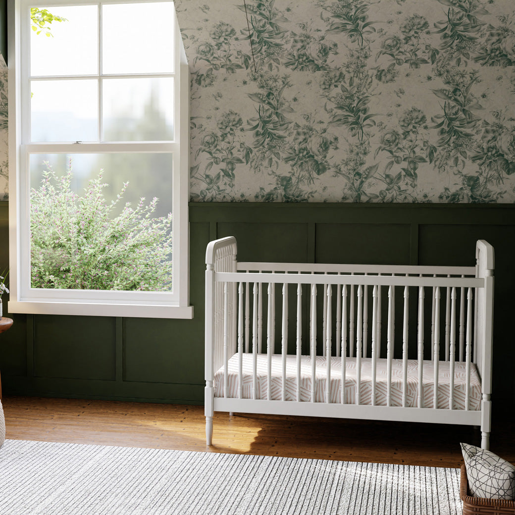 Full lifestyle view of The Namesake Liberty 3-in-1 Convertible Spindle Crib in a green room, next to a window in -- Color_Warm White
