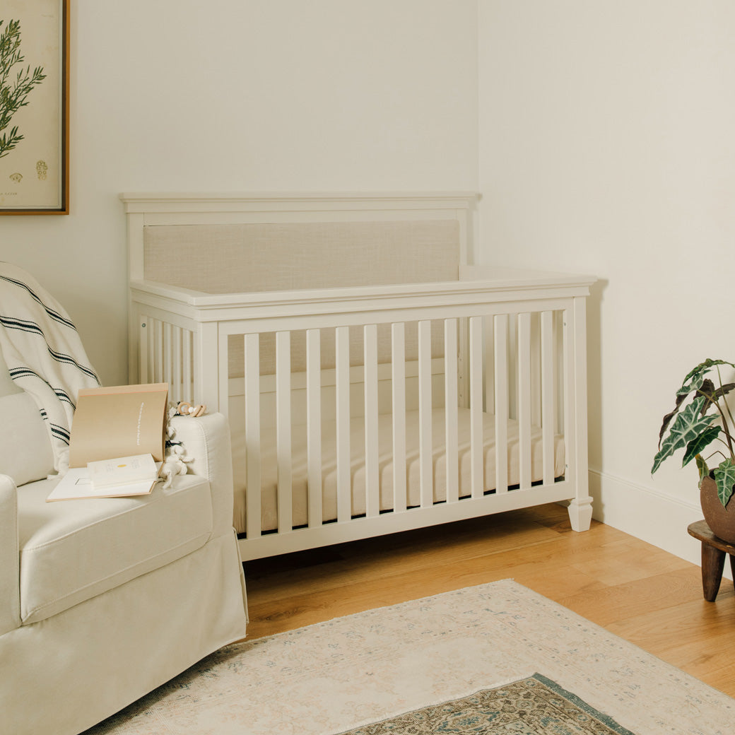 Darlington 4-in-1 Convertible Crib in Warm White next to a recliner 