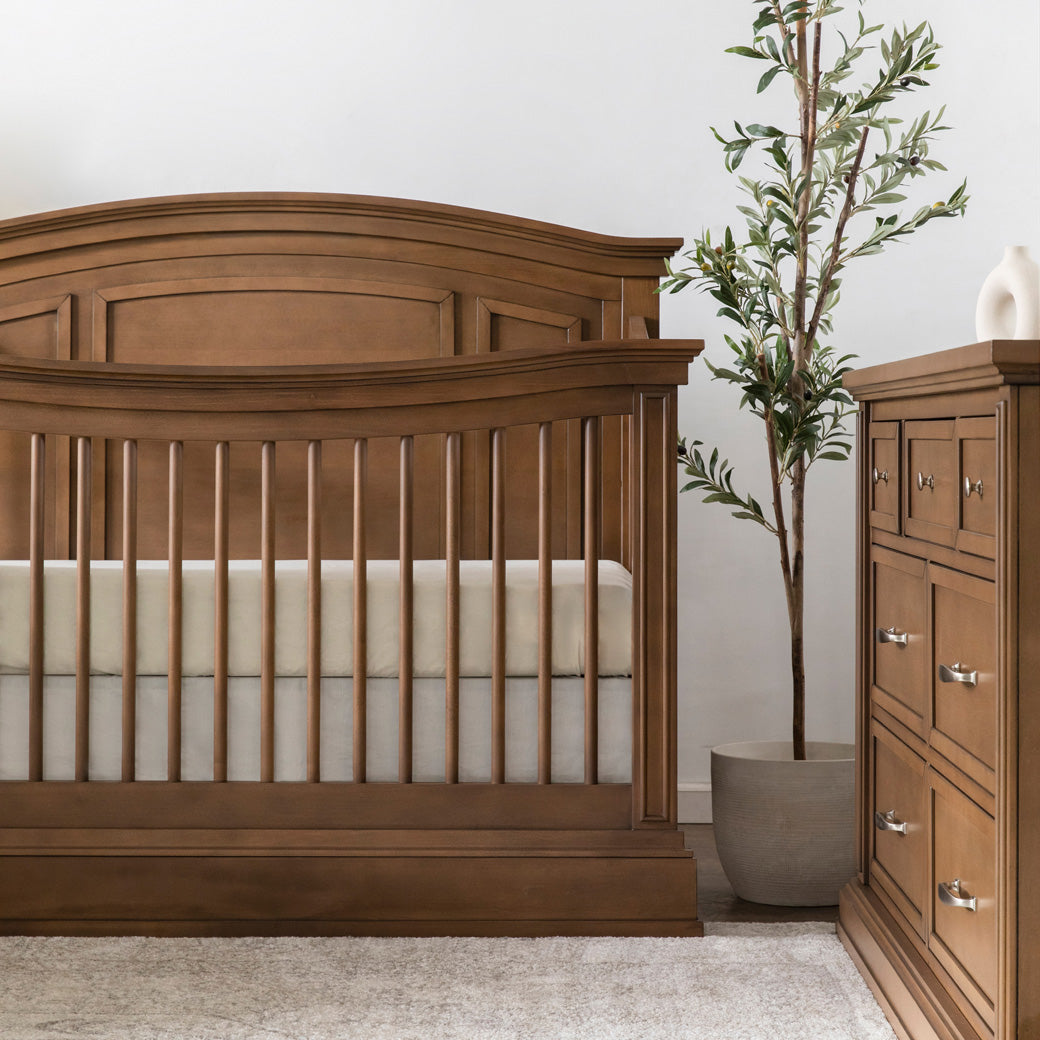 Namesake's Durham 4-in-1 Convertible Crib next to a plant and dresser in -- Color_Derby Brown