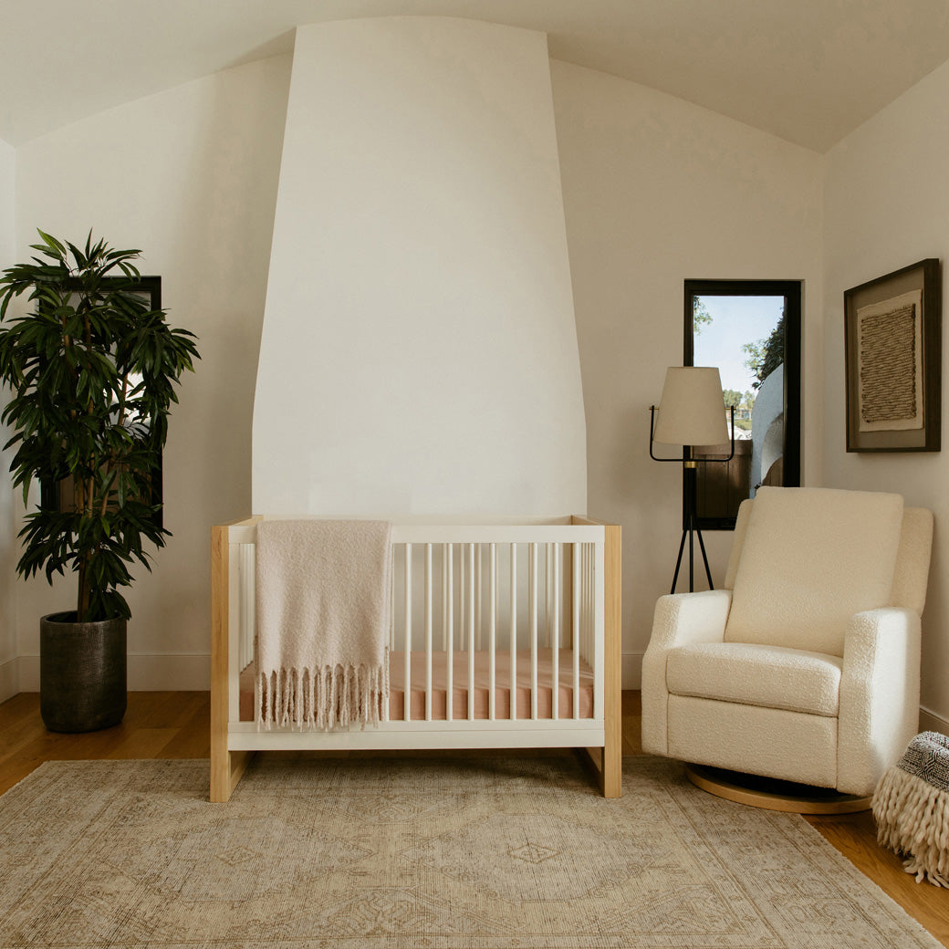 Front view of Namesake's Nantucket 3-in-1 Convertible Crib next to a recliner  in -- Color_Warm White/Honey
