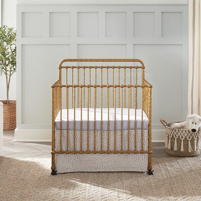 Front view of Namesake's Winston 4-in-1 Convertible Mini Crib next to a basket  in -- Color_Vintage Gold