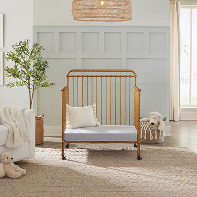 Namesake's Winston 4-in-1 Convertible Mini Crib as daybed next to a basket and recliner  in -- Color_Vintage Gold