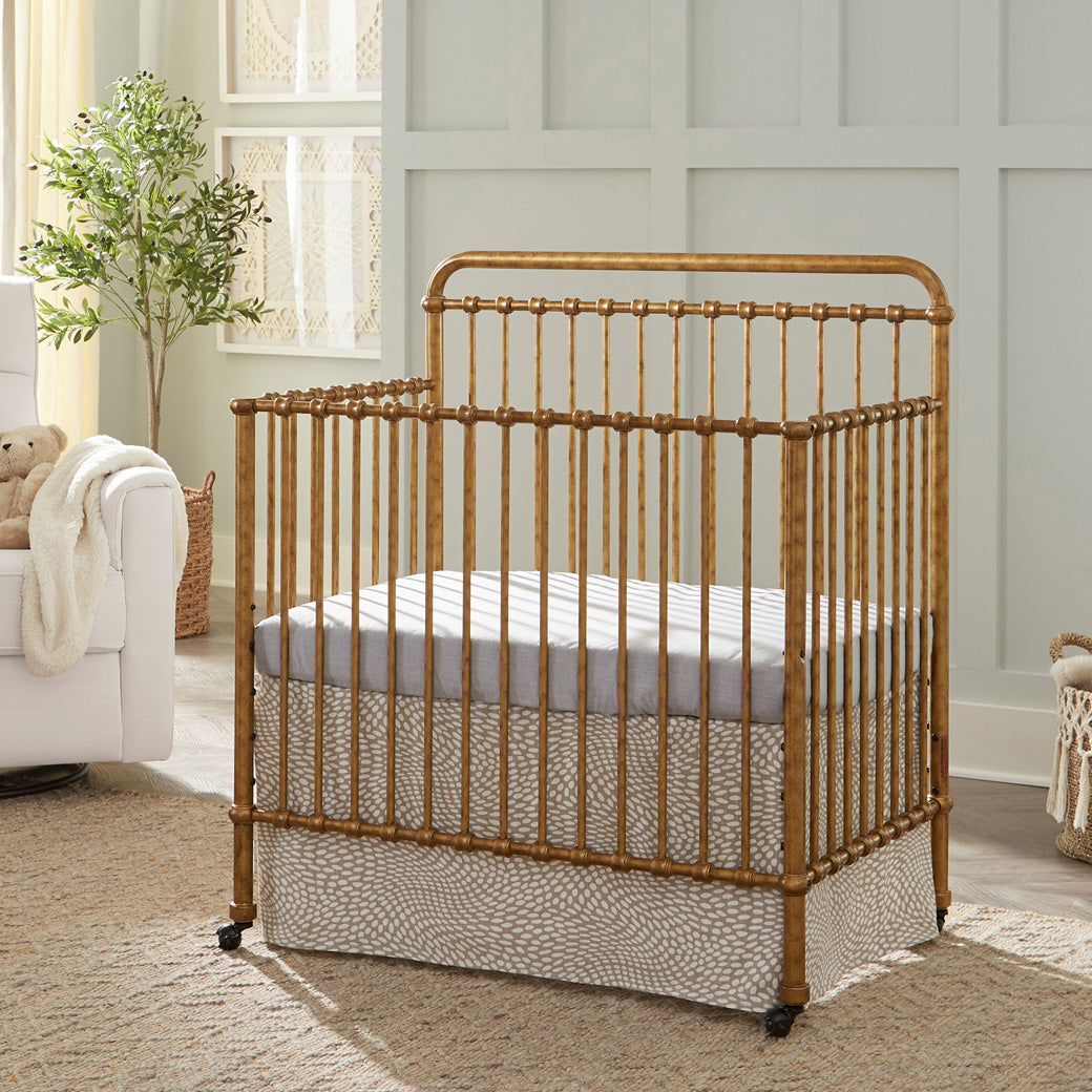 Namesake's Winston 4-in-1 Convertible Mini Crib next to a recliner  in -- Color_Vintage Gold