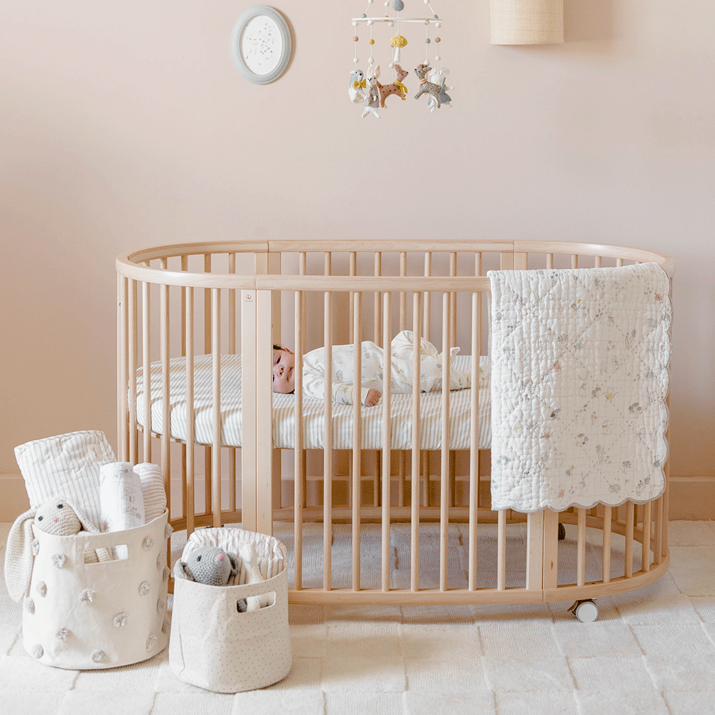 A baby in a crib equipped with a Stokke's Sleepi V3 Fitted Sheet By Pehr in -- Color_Stripes Away Pebbles