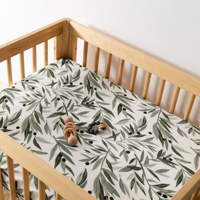 A crib with toy equipped with Babyletto's All-Stages Midi Crib Sheet In GOTS Certified Organic Muslin Cotton in -- Color_Olive Branches