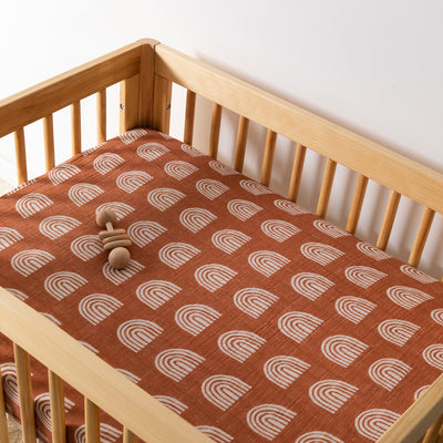 A crib with toy equipped with Babyletto's All-Stages Midi Crib Sheet In GOTS Certified Organic Muslin Cotton in -- Color_Terracotta Rainbow
