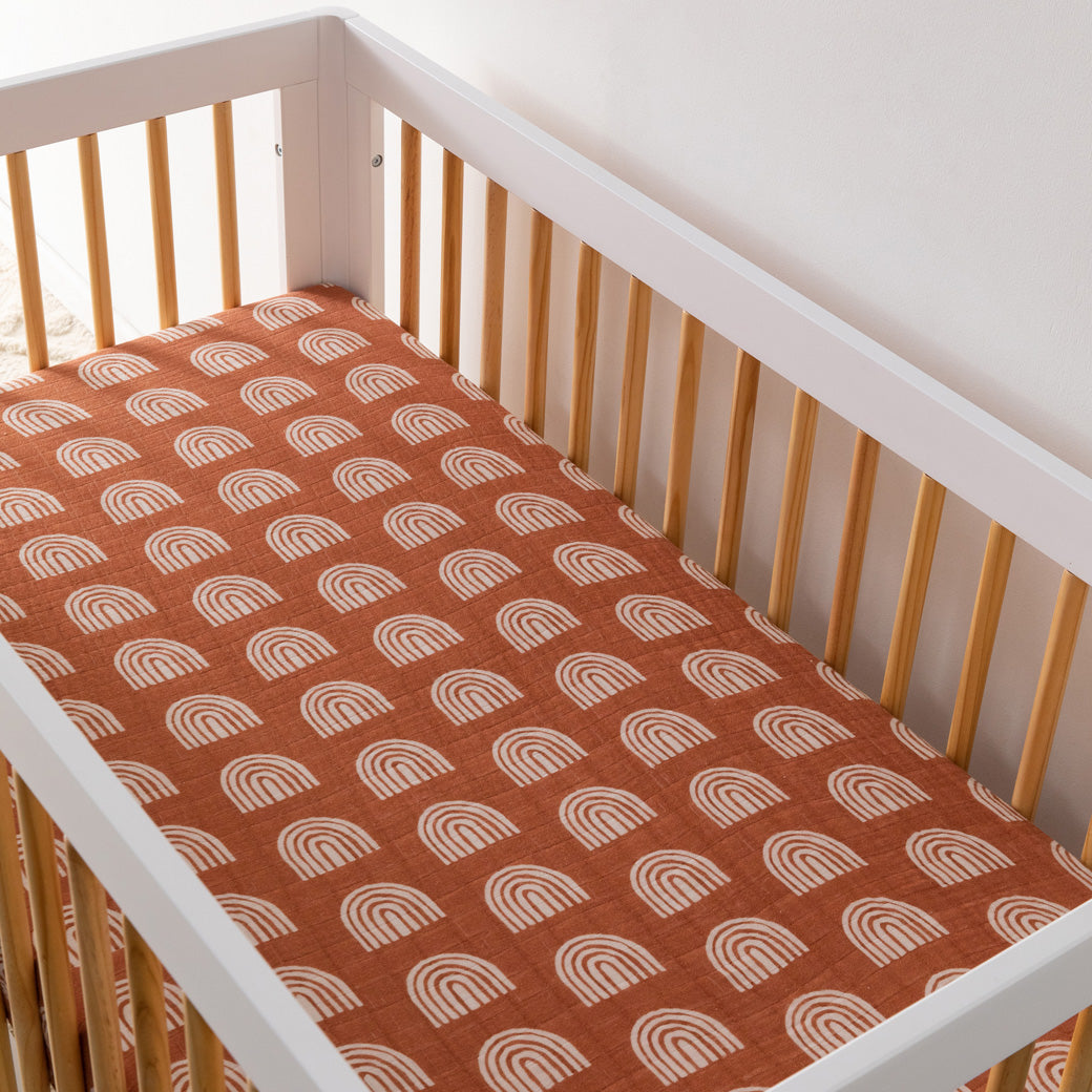 An empty crib equipped with the Babyletto's Crib Sheet in GOTS Certified Organic Muslin Cotton in -- Color_Terracotta Rainbow