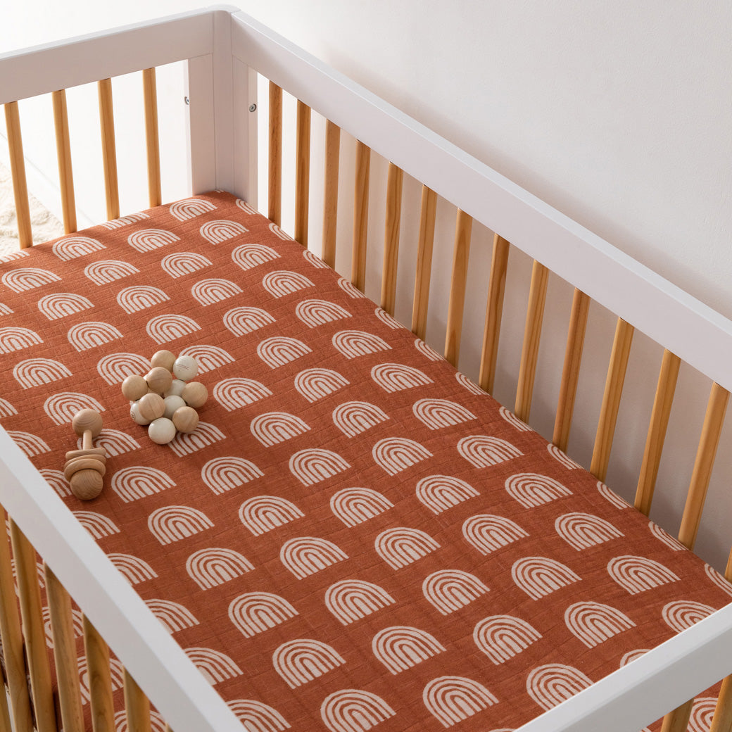 A crib with toy equipped with the Babyletto's Crib Sheet in GOTS Certified Organic Muslin Cotton in -- Color_Terracotta Rainbow