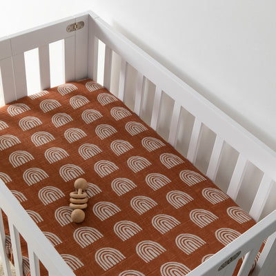 A crib with a toy inside equipped with Babyletto's Mini Crib Sheet in -- Color_Terracotta Rainbow