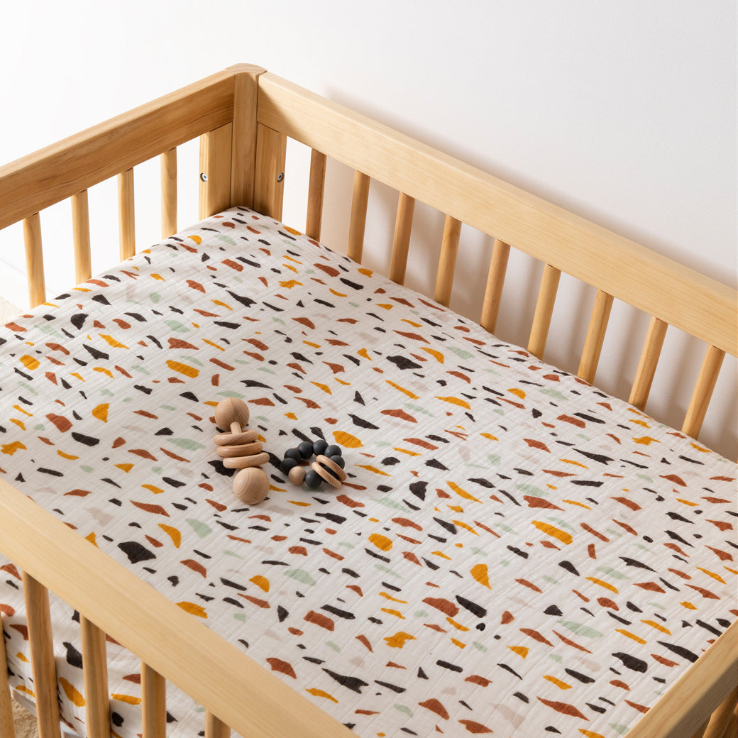 A crib with toy equipped with Babyletto's All-Stages Midi Crib Sheet In GOTS Certified Organic Muslin Cotton in -- Color_Terrazzo