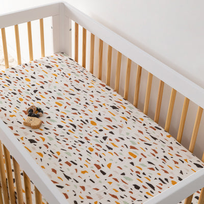 A crib with toy equipped with the Babyletto's Crib Sheet in GOTS Certified Organic Muslin Cotton in -- Color_Terrazzo