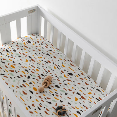 A crib with a toy inside equipped with Babyletto's Mini Crib Sheet in -- Color_Terrazzo